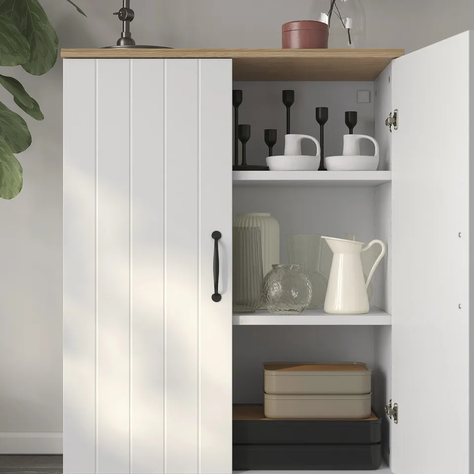 A white cabinet with grooved doors and wooden top, housing decorative items on its shelves.