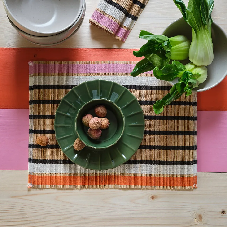 A woven place mat with banana fibres and cotton. It protects a tabletop and reduces noise from dishes. Green bowl with lychees sits on it.