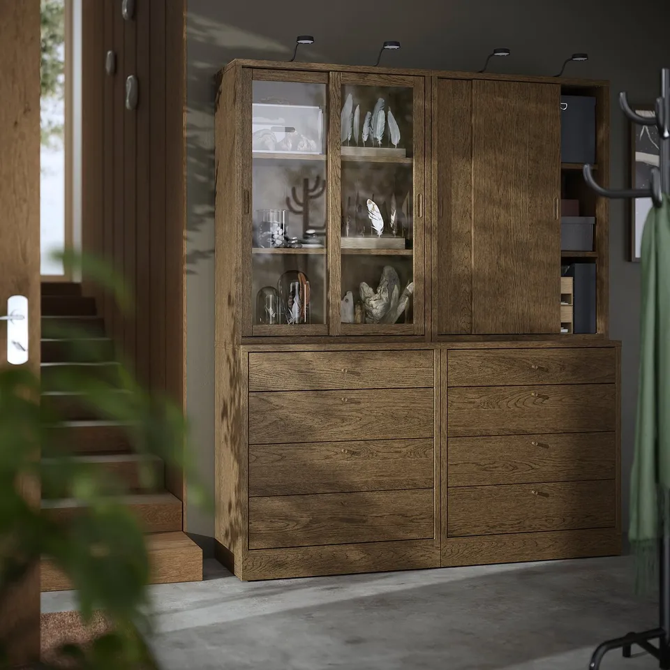 Brown wooden TONSTAD cabinet with glass doors houses decor, sits between tall plants and stairs.