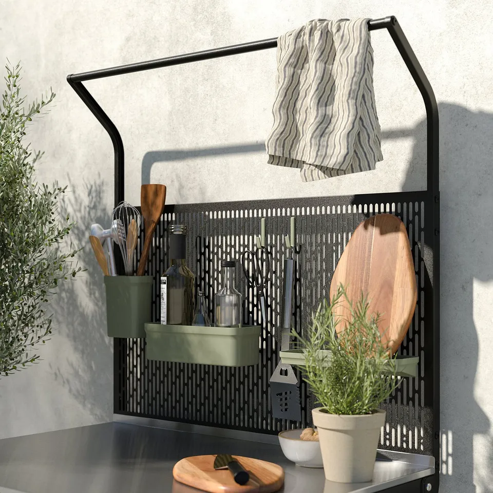 Black metal kitchen shelf with green bins, utensils, and striped towel, stainless steel countertop, wooden cutting board, and potted plant.