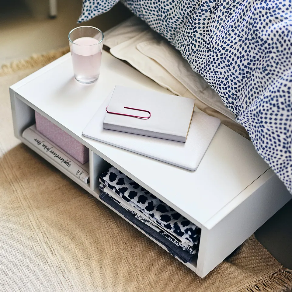 White modern side table with open shelves and white items on top, next to a bed with blue patterned bedding, on a beige rug.