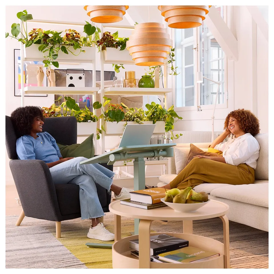 Two people chat in a bright room; one in blue shirt, the other in patterned pants. A light wooden laptop table with books and fruit.