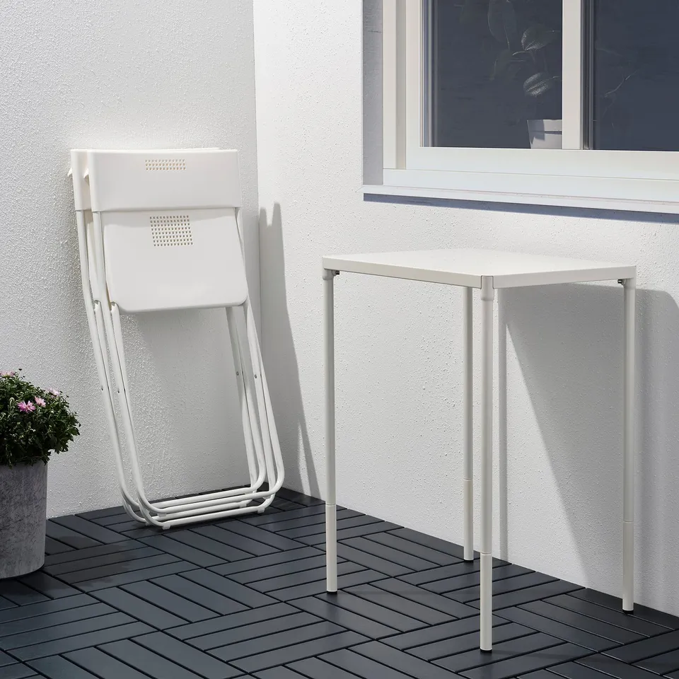 Two white metal chairs, one folded, placed near a small white square table on a black tiled patio.