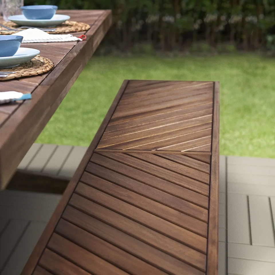 NORRMANSÖ garden bench in acacia wood with herringbone pattern, placed on lawn near dinner table.
