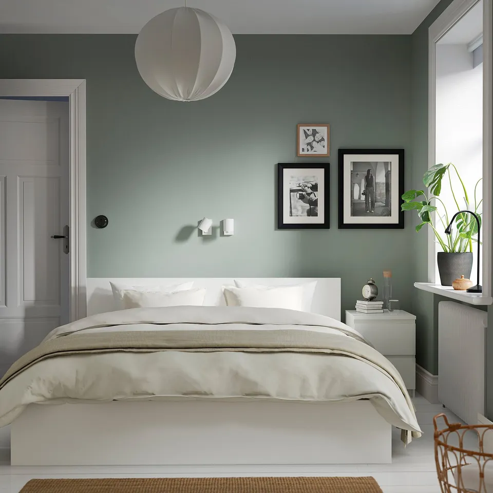 Minimalist bedroom with MALM bed, sage-green walls, bedside table with books, plant, candle, and wicker chair. Wooden floors, calm atmosphere.