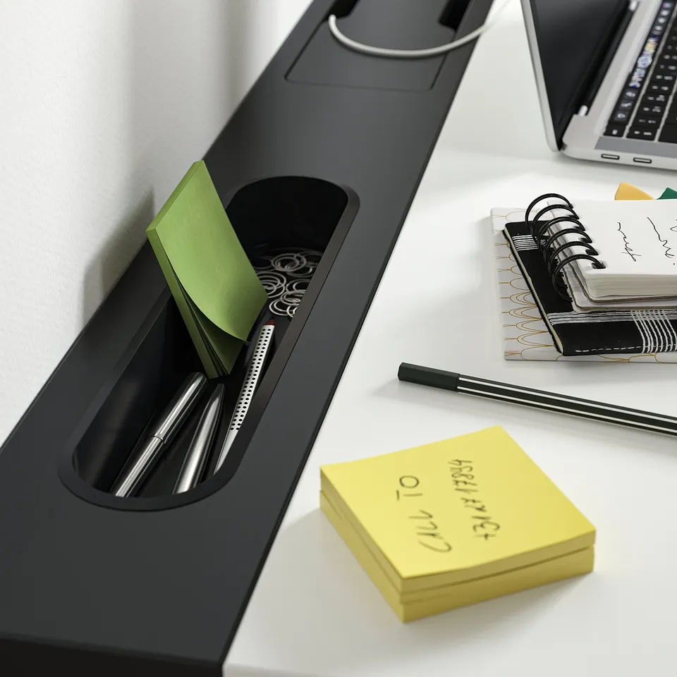 A black desk organiser with pens, notebooks, and sticky notes on a white desk, alongside a laptop.