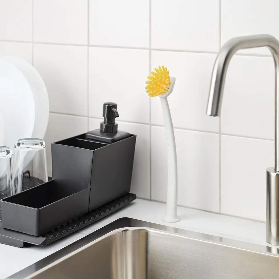 Black kitchen sink organiser with soap dispenser and yellow brush, featuring a scraper edge for removing food debris.