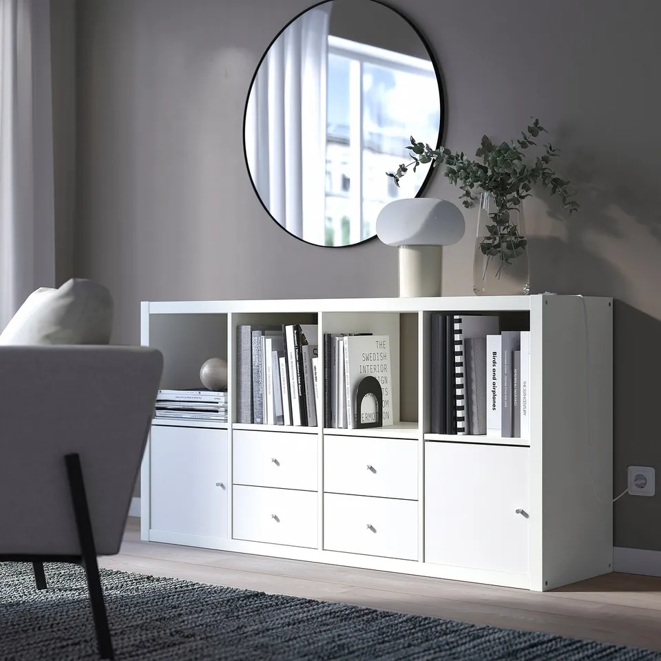 KALLAX storage unit with shelves and drawers, holding books and a vase, against a grey wall in a minimalist living space.