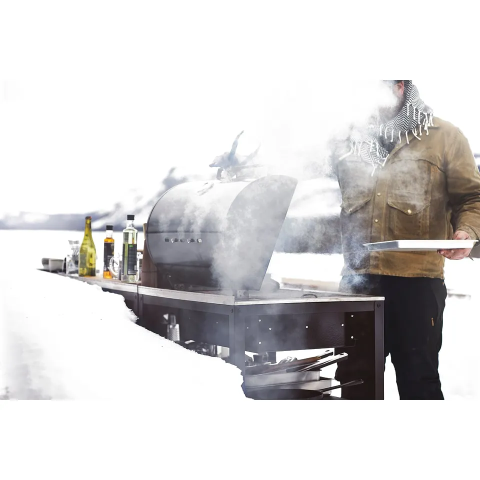 Person grilling in snowy outdoor setting, using a stainless steel GRILLSKÄR charcoal grill. Steam visible, surrounded by bottles.