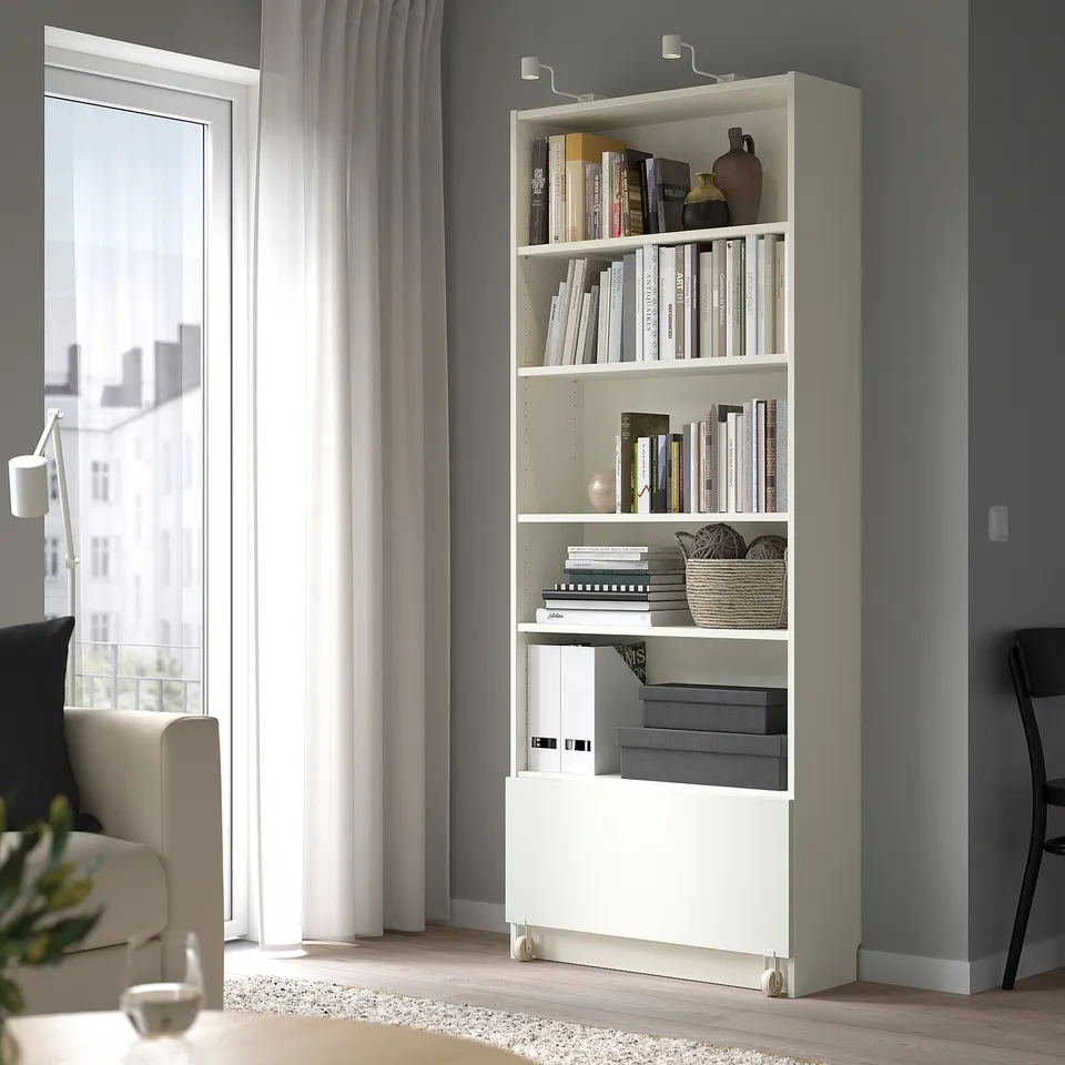 White BILLY bookcase with books, decor, drawer, near window, couch, & table.