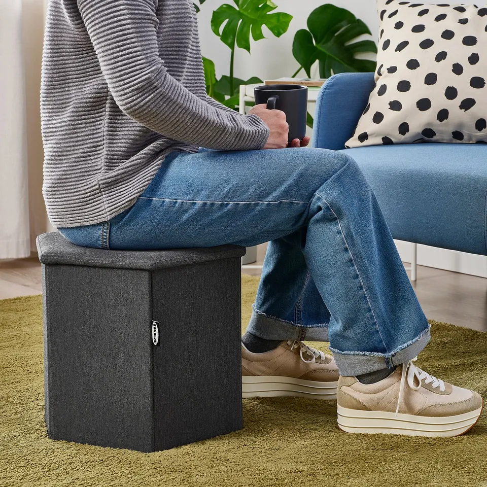Person sitting on blue couch, resting foot on grey KJUGE storage stool, holding coffee cup.