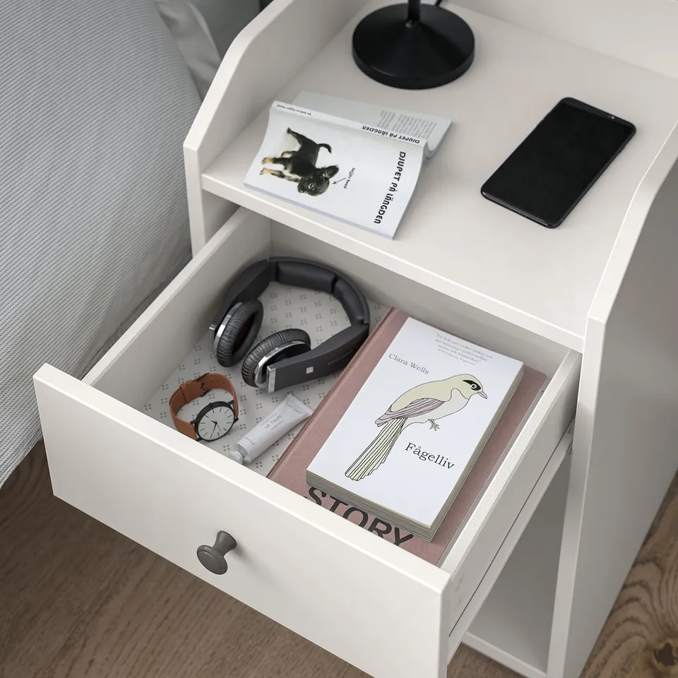 White bedside table with drawer open, showing headphones, book story, and a phone on top.