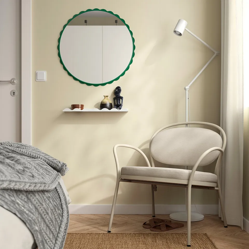 Round mirror with green frame on minimalist wall above white shelf with decor, contrasting beige chair below.
