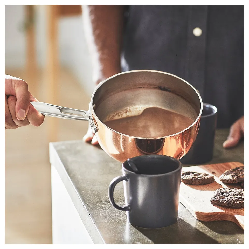 FINMAT copper saucepan with steel handle is held over a blue mug and chocolate treats on a wooden board. Modern kitchen setting.