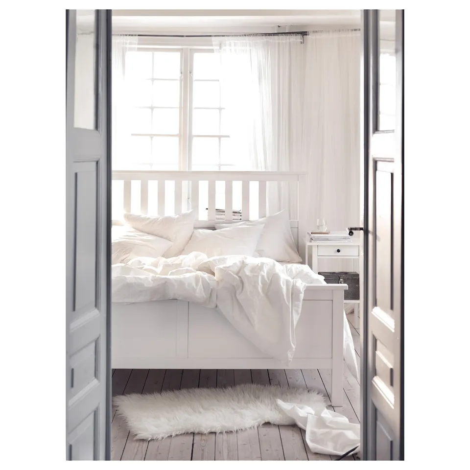 Minimalist white wooden bed with high headboard, white bedding, plush rug, and bedside table with lamp.