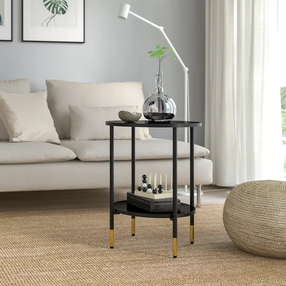 Modern black metal side table with gold accents and glass shelves, beside sofa with vase, plant, and candles.