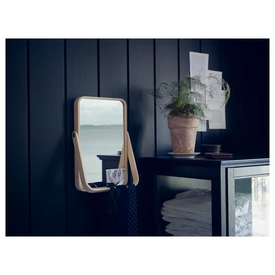 IKORNNES mirror in wood frame on dark cabinet, reflects plant and notes, with tie hanging.