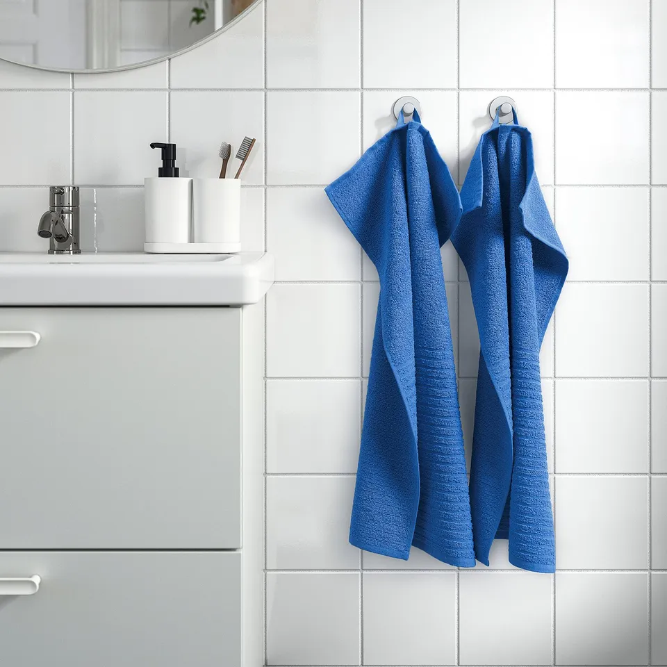 Blue VÅGSJÖN towels hang on bathroom wall hooks, next to sink with mirror, soap dispenser, and toothbrushes.