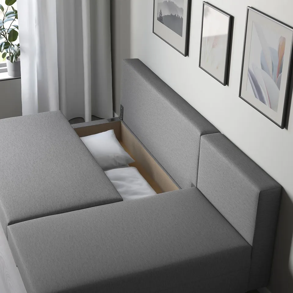 A modern grey sofa with a hidden bed compartment open, displaying two white pillows and wooden base. Ideal for space-saving.