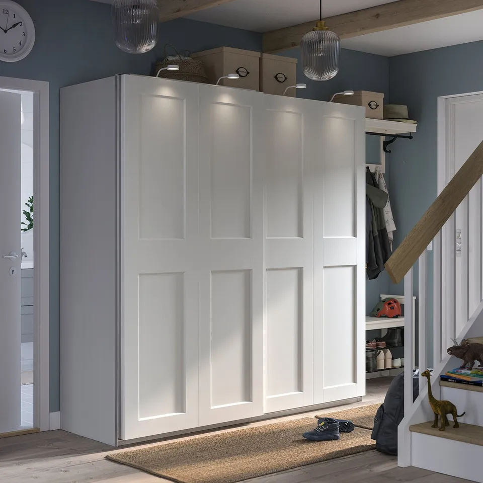 Modern white PAX wardrobe with sliding doors, featuring integrated lighting and mesh basket, set against a blue wall.