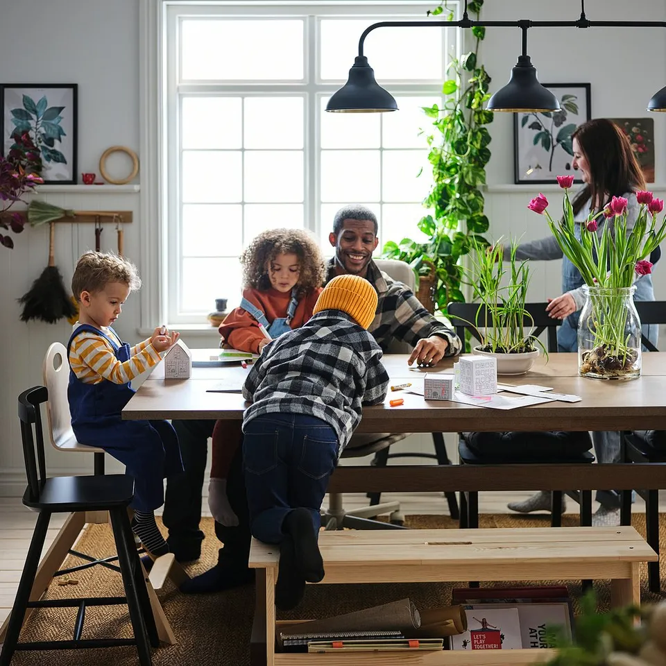 Family crafts together at wooden table with lights and plants.