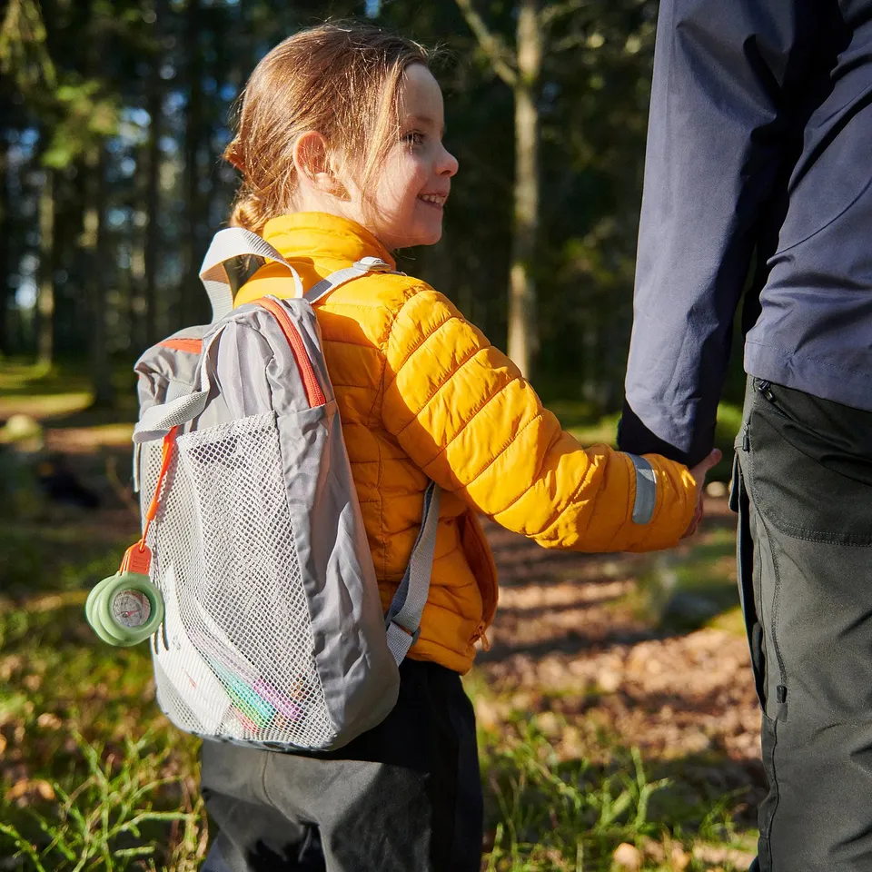 Child wearing yellow jacket and grey backpack outdoors. Backpack has a mesh pocket with compass and magnifying glass. Adult stands beside smiling.