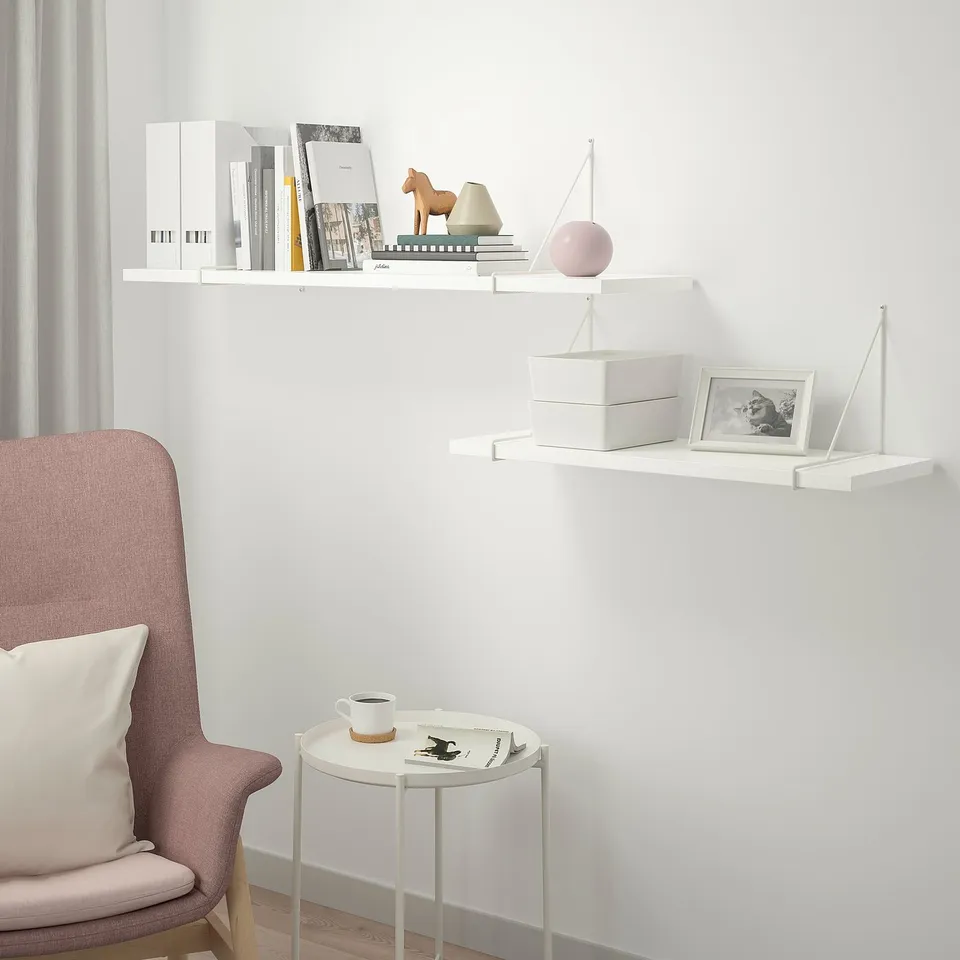 White BERGSHULT shelf with bevelled and smooth edges, holding various items. Pink chair and small table, with coffee cup and magazines.