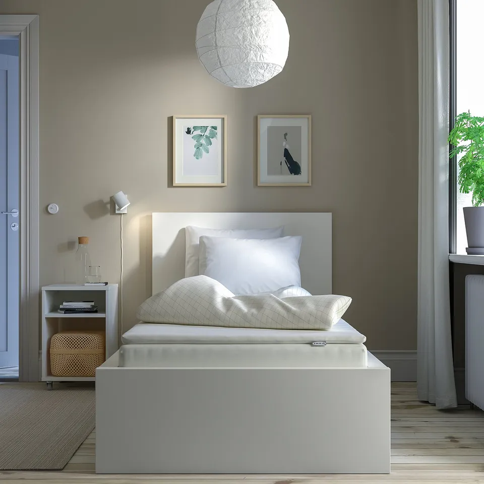 Minimalist bedroom with NÄSFJÄLLET white bed, small bedside table, plant, and two wall pictures. Grey walls and wooden floors.