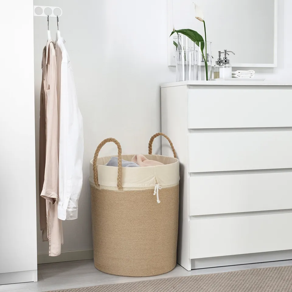 LJUNGAN beige jute laundry basket, large, with handles, near white dresser, holding clothes.