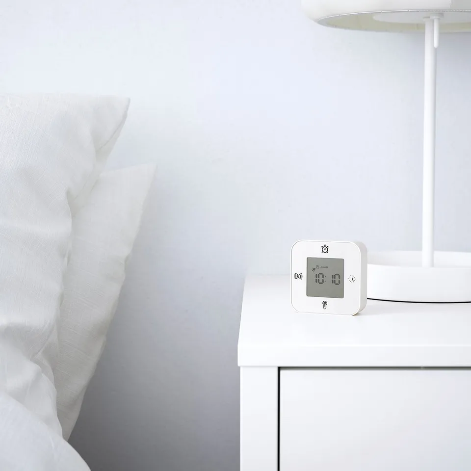 A modern bedside alarm clock displaying time on a white bedside table with a lamp and crisp white bedding.
