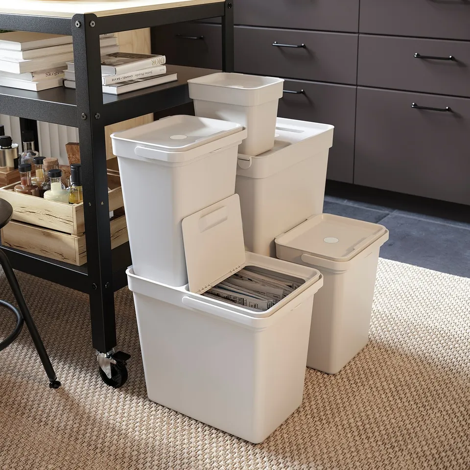 White HÅLLBAR recycling bins, three sizes stacked; two open, one closed, near black shelf with books and wooden boxes.