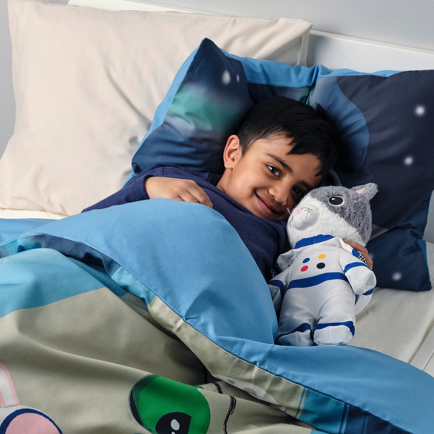 Happy child lying on bed, hugging blue and white astronaut cat cuddly toy, using star-patterned pillow.
