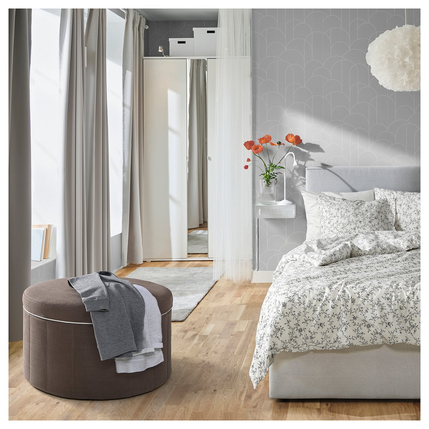 Modern bedroom: light floors, floral bed, footstool, large windows with grey curtains.