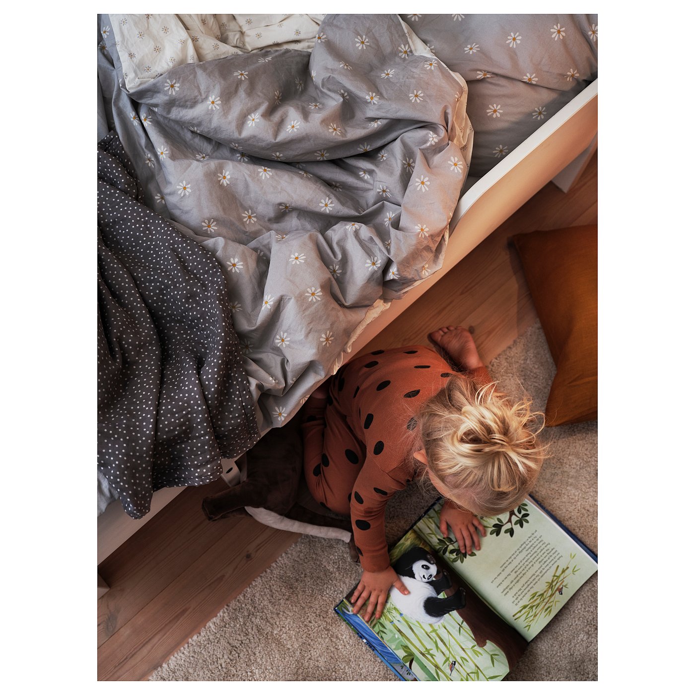 A child reads under a bed dressed with a grey and white daisy-patterned NATTSLÄNDA duvet. The child wears a spotted, orange onesie.