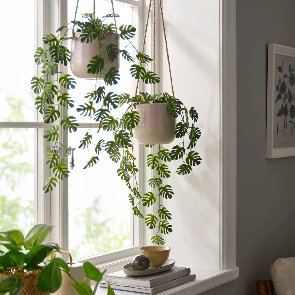 FEJKA hanging plants in white pots with green leaves by window, complementing books and bowl.
