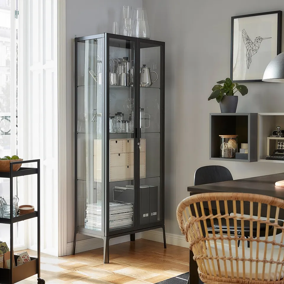 Black MILSBO glass-door cabinet displays glassware, secured with a lock. It adjustable shelves and a cable outlet.