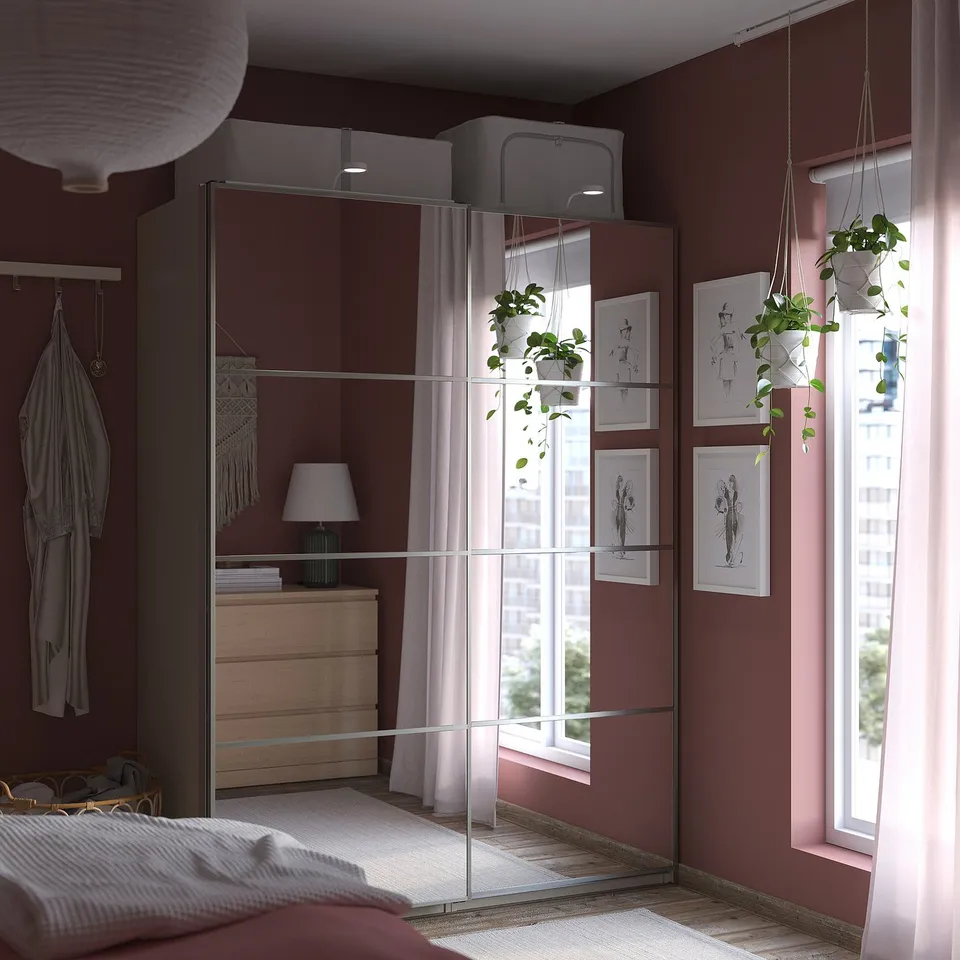 Modern bedroom with burgundy walls, featuring a reflective PAX wardrobe, hanging plants, and a window with sheer curtains.