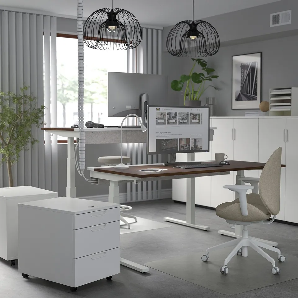 Modern home office with adjustable desk, chair, pendant lights, cabinets, and large monitor.