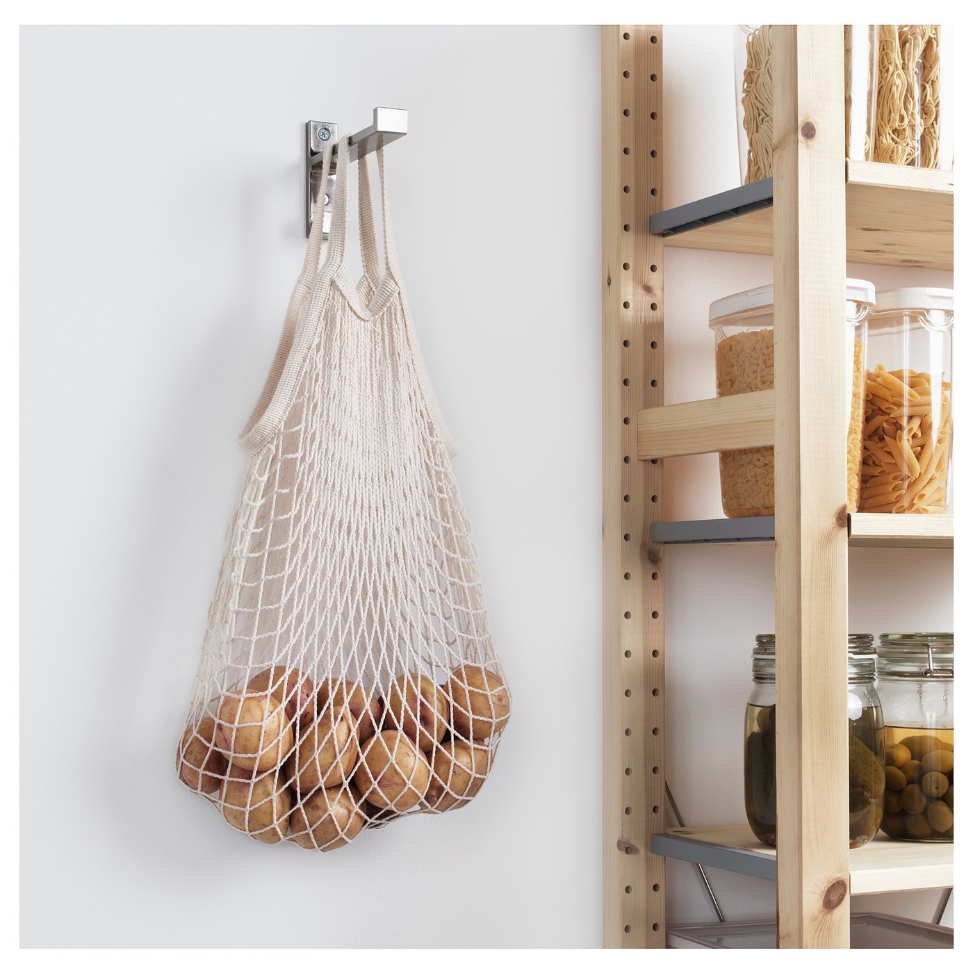 Mesh produce bags hang on wall hook in kitchen, storing potatoes beside wooden shelving units with food items.