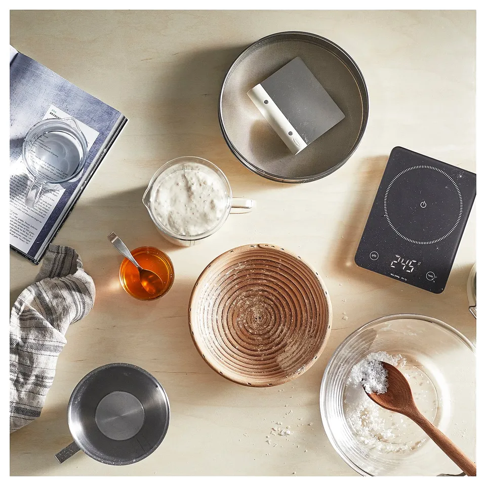 Top view of baking tools: GENOMSNITT device, bowls, utensils, cloth, and recipe book on a counter.