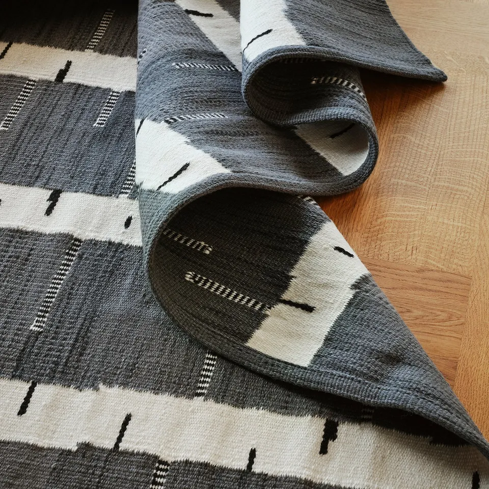 Roll-out handwoven wool rug, grey with birch pattern, laid out on wooden floor. High-quality, reversible, and durable.