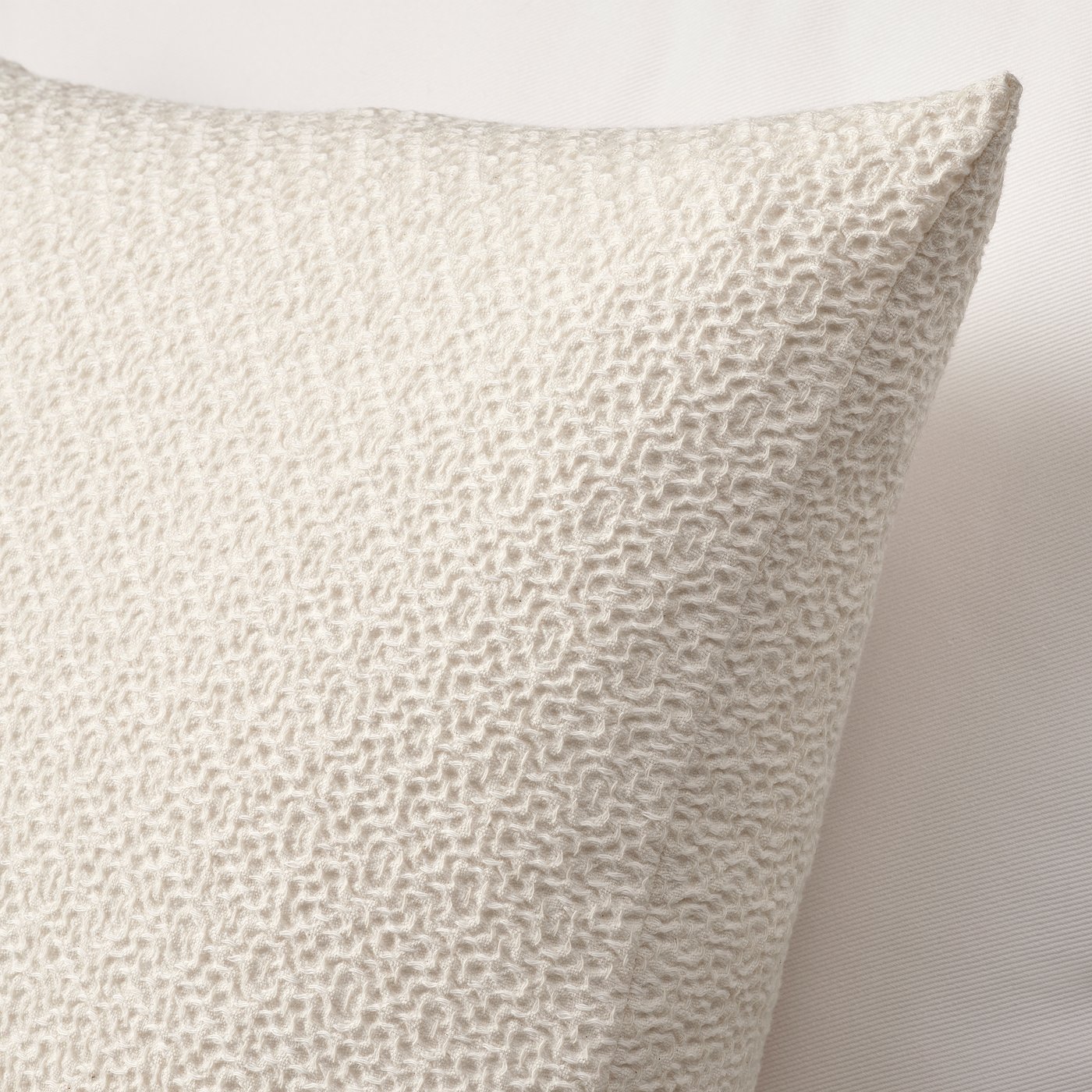 Soft, textured beige cotton cushion cover, showing intricate waffle pattern against light wall. Zip at one edge.