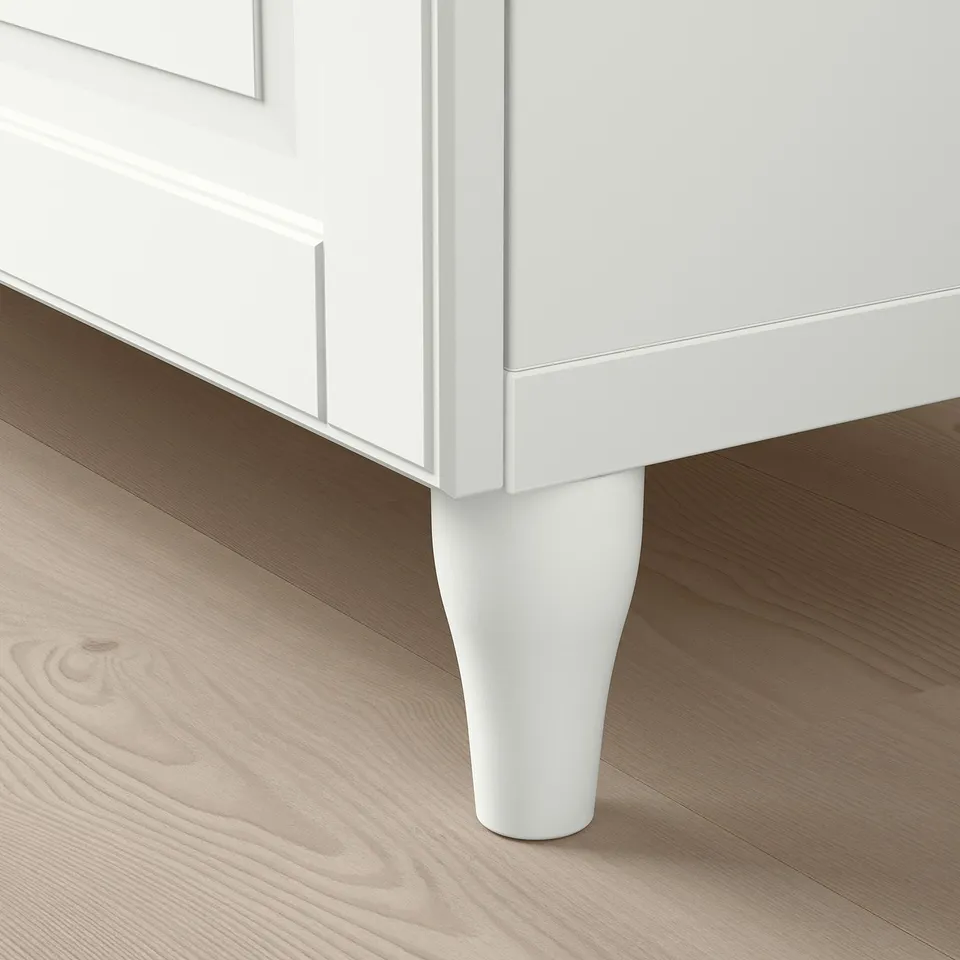 Modern white cabinet with tall, tapered leg on light wooden floor.