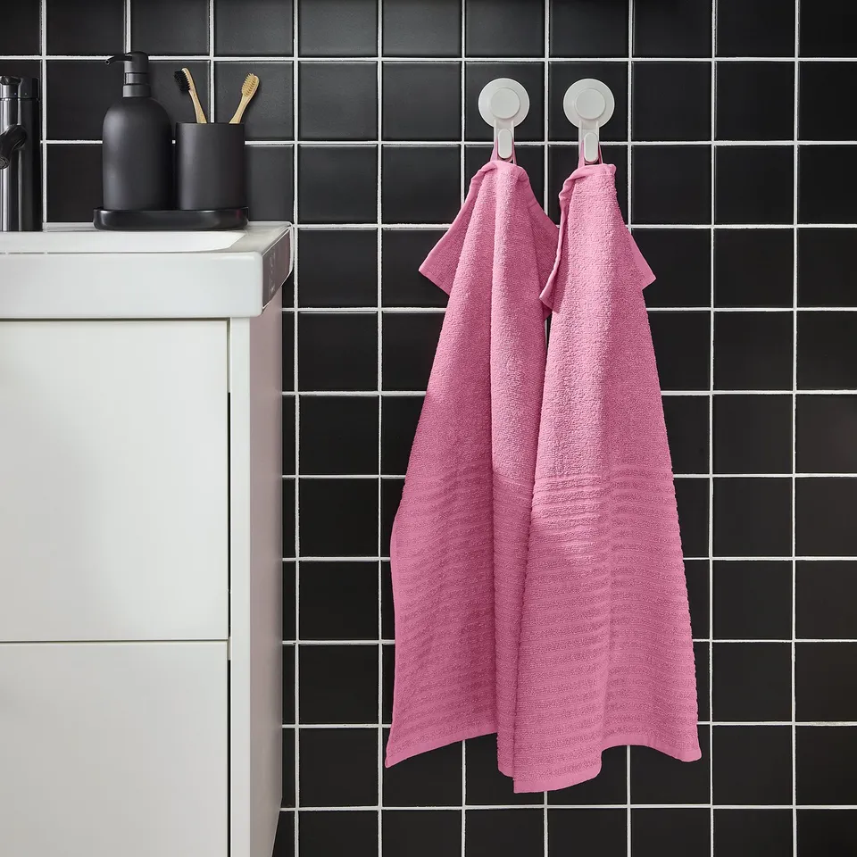 Two pink VÅGSJÖN towels hang on hooks next to a black-tiled wall and white cabinet, adding colour to the bathroom.