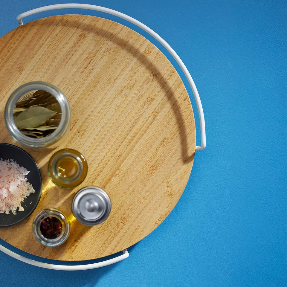 Wooden turntable with raised edges on blue surface, holding spices, oil, and leaves. Keeps items organised and easily accessible.