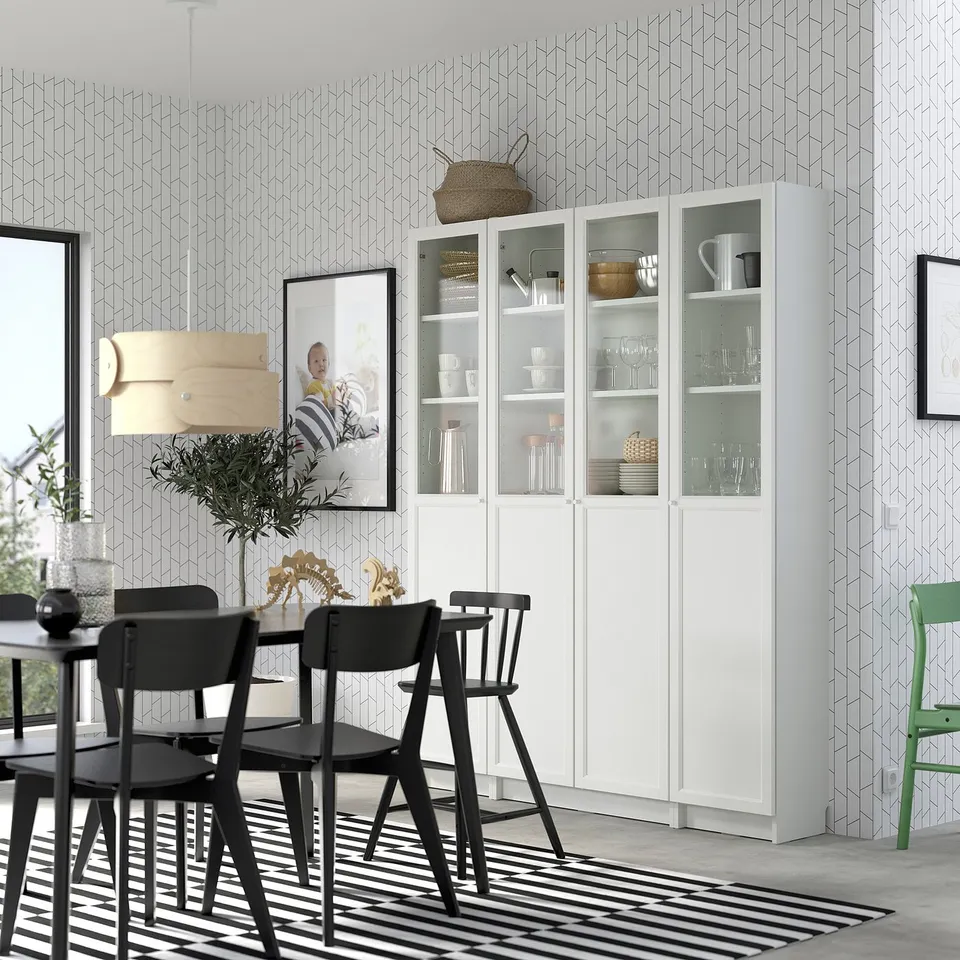Modern dining room with white BILLY bookcase, black chairs, geometric wallpaper, striped rug, and potted plant.