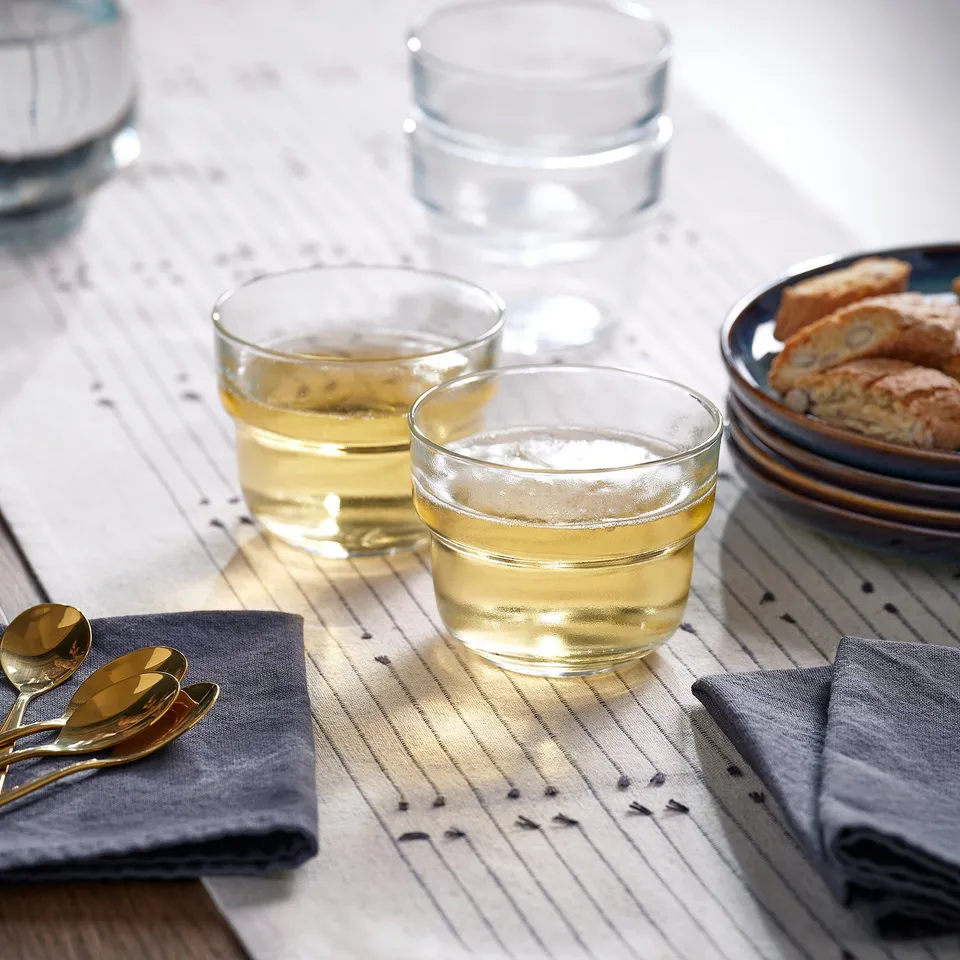 Two glass cups on a striped tablecloth, filled with a yellow liquid, placed beside grey napkins and gold spoons; one cup has a white substance on top.