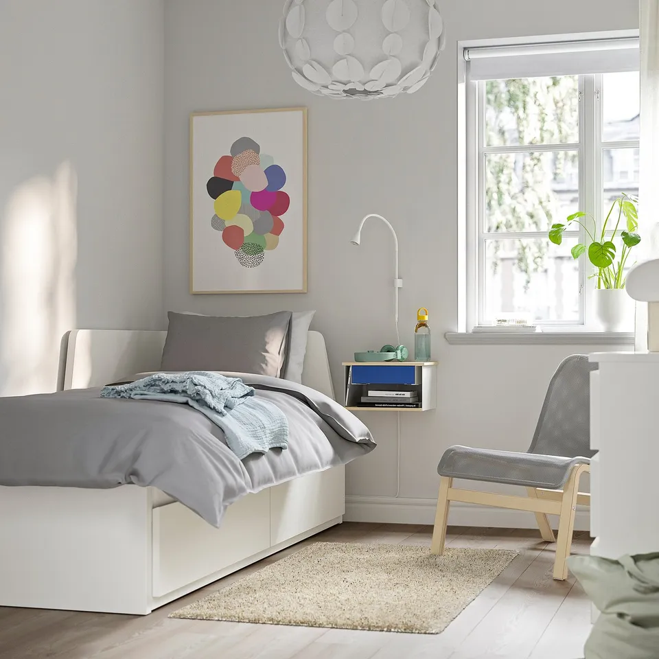 Modern bedroom with white bed, STOMSÖ blue bedside table, colourful wall art, white pendant light, and grey chair.