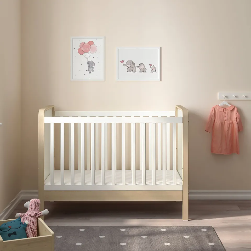 A nursery featuring a white ÄLSKVÄRD cot, grey rug, elephant art, orange dress, and toy basket in a minimalistic, beige space.