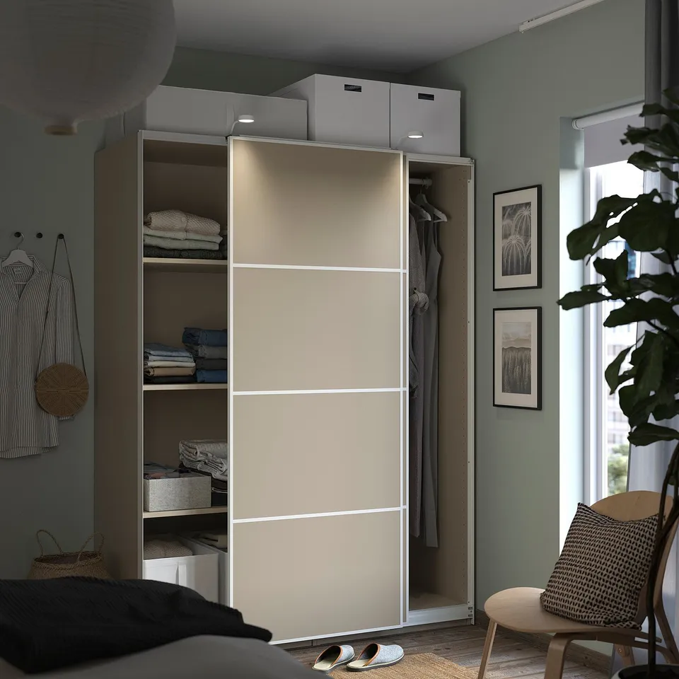 Light beige wardrobe with sliding doors, white shelves, clothes, and storage in a green-walled bedroom with a plant.